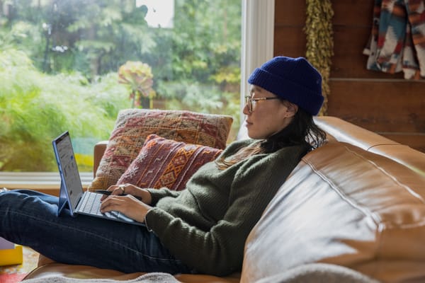 A person in jeans, a green sweater, blue hat and glasses sits on a brown couch by a window, with a laptop on their legs